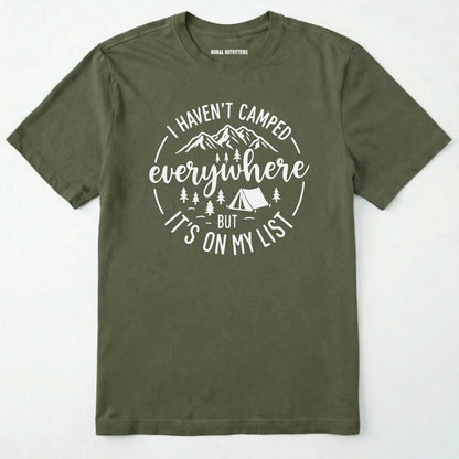 Military Green funny camping shirt saying with mountains, trees, tent graphic and "I Haven't Camped Everywhere But It's On My List" text on a white background.