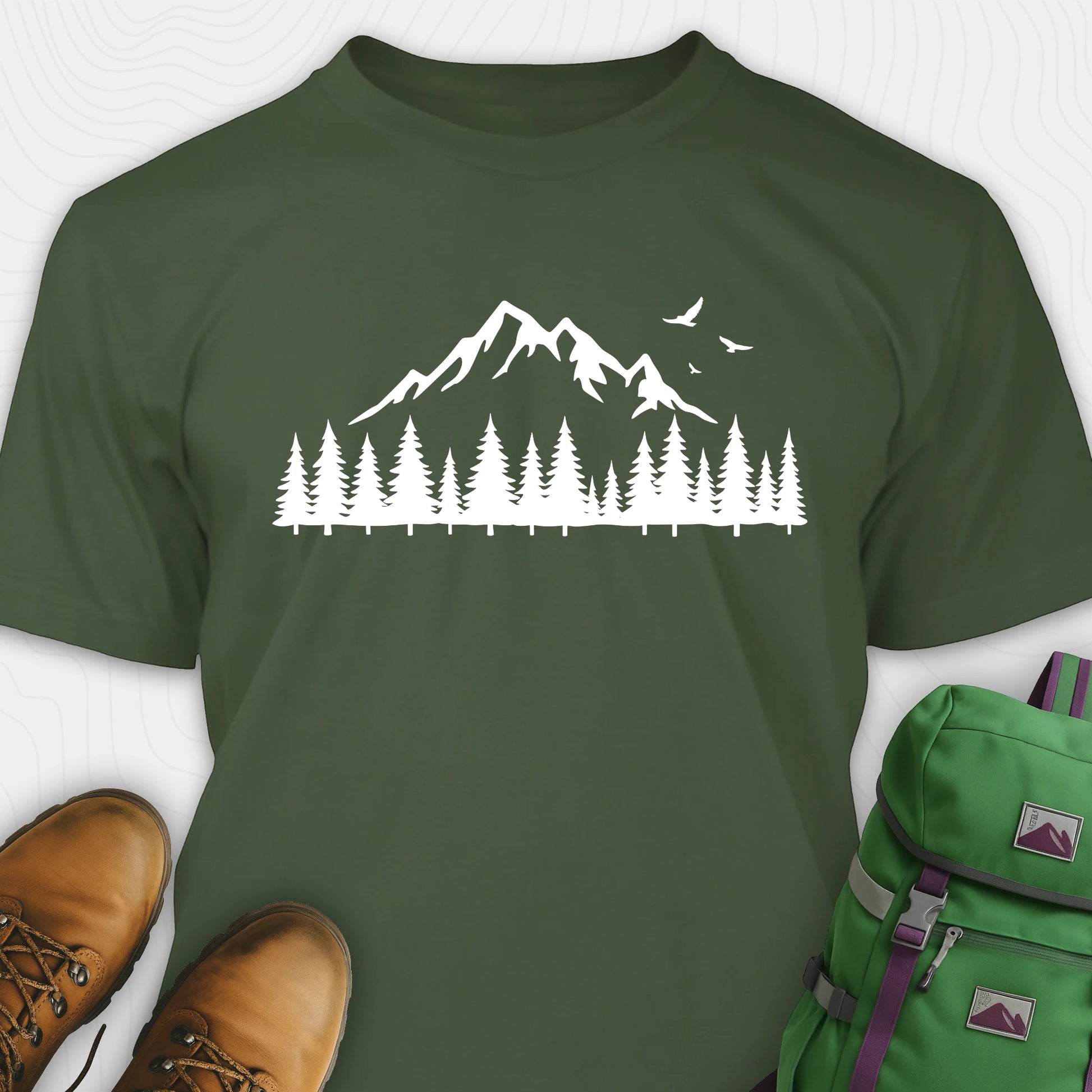 Green wild t-shirt with a minimalist mountain and pine tree graphic, shown flat with boots and a backpack
