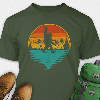 Military green bigfoot shirt for men with retro mountain graphic beside boots and green backpack