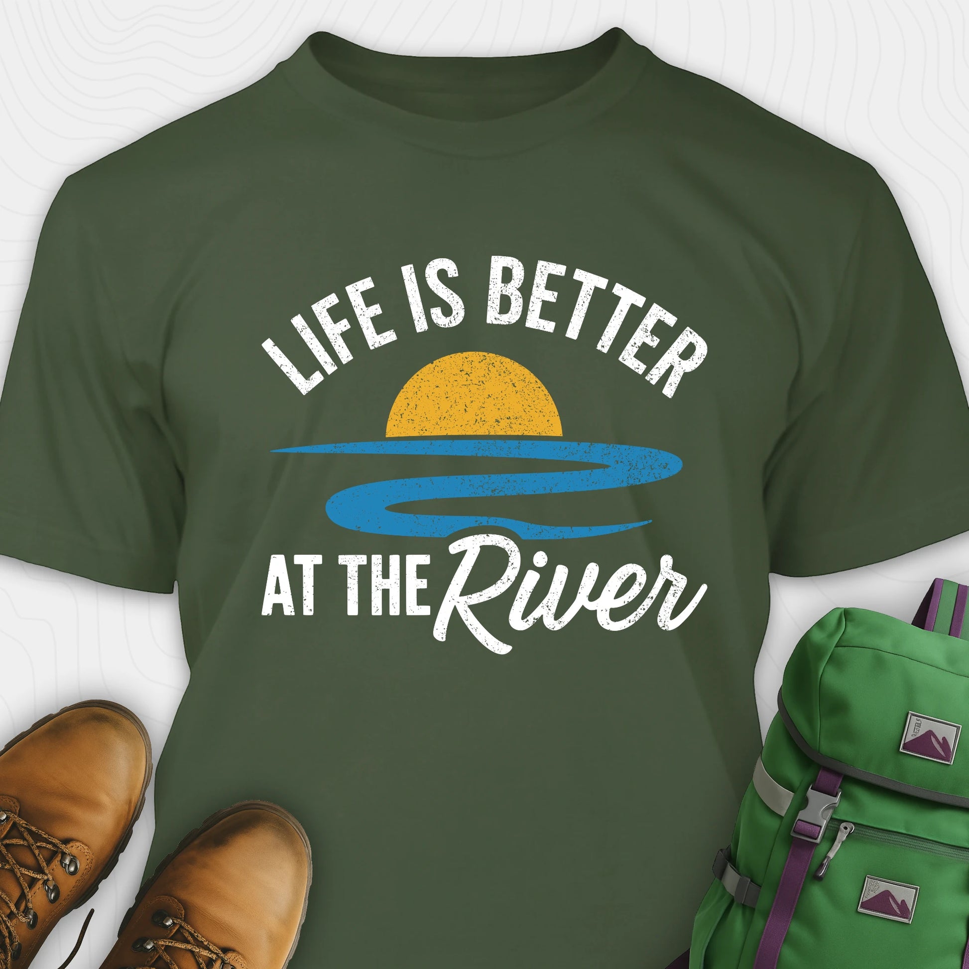 Green river shirt with 'Life is Better at the River' design, paired with brown boots and a green backpack on a light gray background.