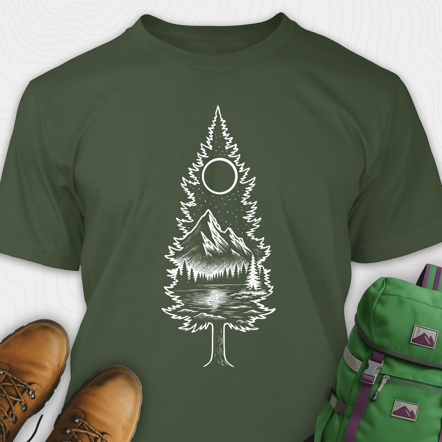 A green tree t-shirt featuring a detailed white graphic of a pine tree silhouette. The tree outline contains a landscape scene: a mountain peak rising over a forested shoreline, with its reflection visible in a lake below. Above the mountain is a starry night sky and a full or partial circle representing the moon or sun. The t-shirt is displayed flat with brown leather boots to the lower left and a green and purple backpack to the lower right, suggesting an outdoor or hiking theme.
