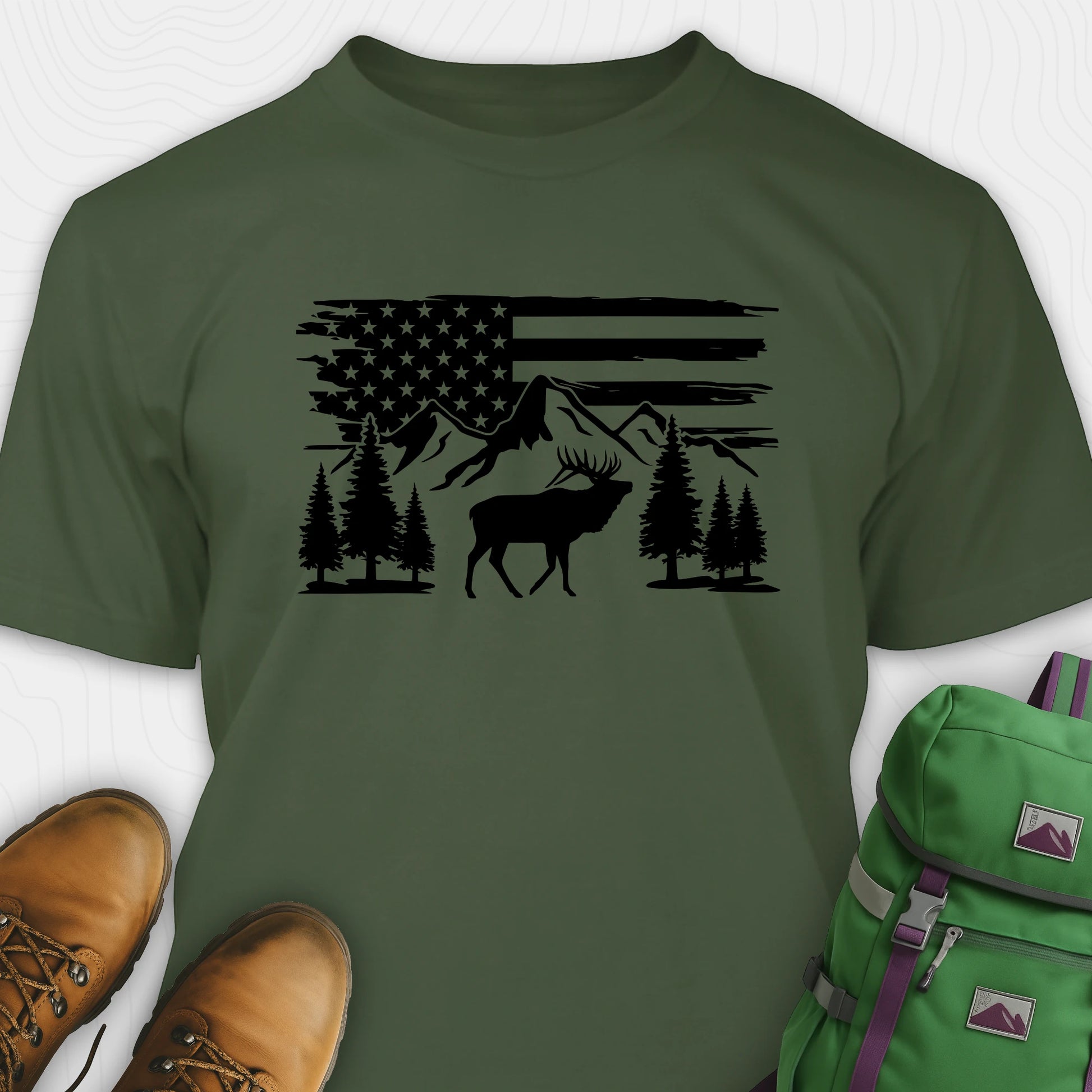 Green deer t-shirt with black graphic design of an American flag, trees, and a moose, accompanied by brown boots and a green backpack.