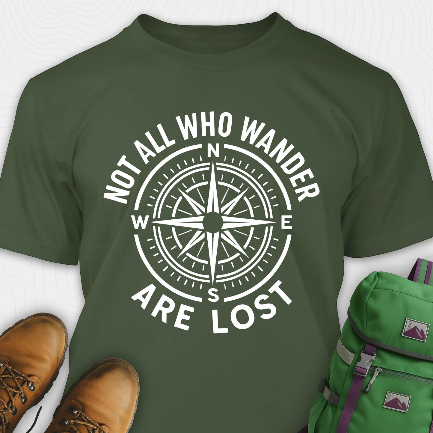 Military green Not All Who Wander Are Lost T-shirt with compass design and text, paired with brown boots and a green backpack on a white background.