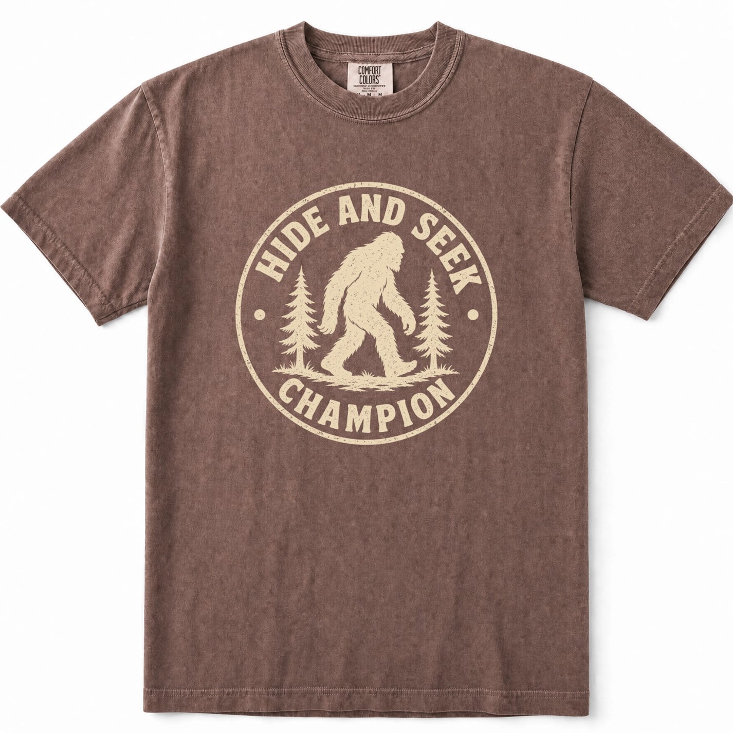 Espresso short-sleeve crewneck t-shirt with a distressed beige graphic on the front that reads 'HIDE AND SEEK CHAMPION' around a Bigfoot silhouette and pine trees.