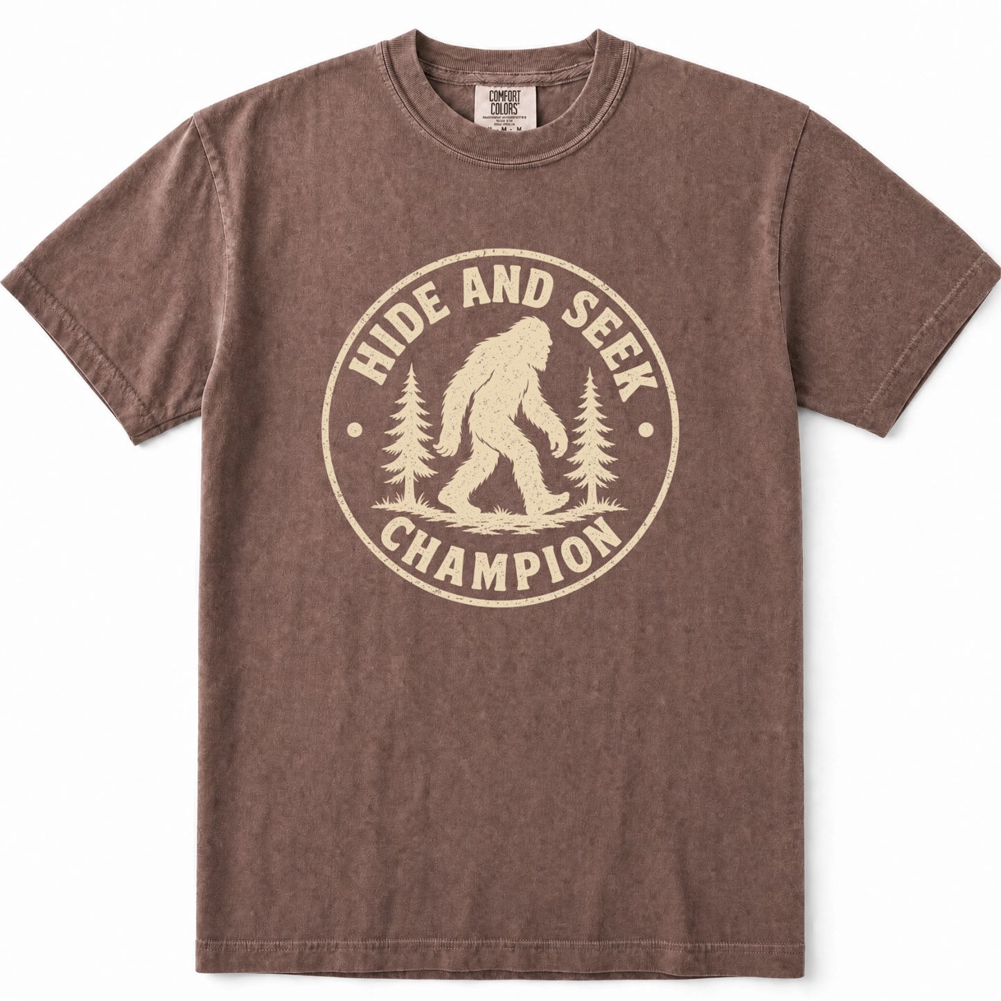 Espresso short-sleeve crewneck t-shirt with a distressed beige graphic on the front that reads 'HIDE AND SEEK CHAMPION' around a Bigfoot silhouette and pine trees.