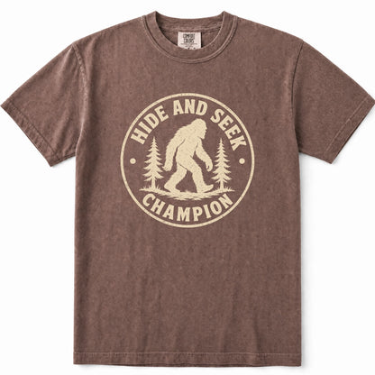 Espresso short-sleeve crewneck t-shirt with a distressed beige graphic on the front that reads 'HIDE AND SEEK CHAMPION' around a Bigfoot silhouette and pine trees.