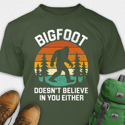 Green funny Bigfoot shirt with sasquatch silhouette and humorous saying, paired with brown boots and a green backpack on a white background.