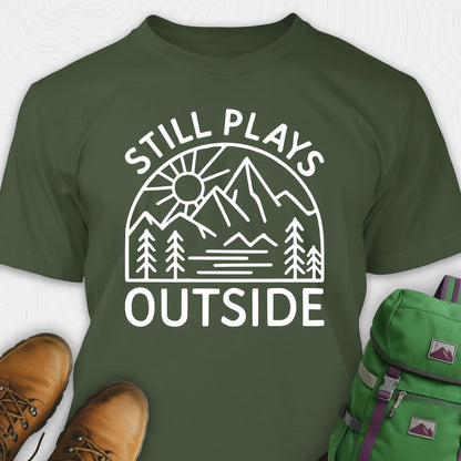 Green t-shirt with 'Still Plays Outside' graphic design, paired with brown boots and a green backpack on a light gray background.
