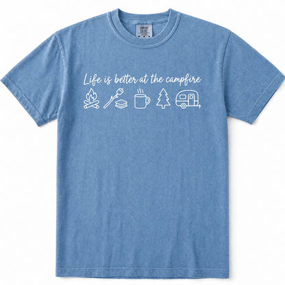 Blue Jean crewneck t-shirt with a white graphic print that reads 'Life is better at the campfire' above simple icons of a campfire, roasting stick, mug, tree, and camper trailer, shown on a plain white background.