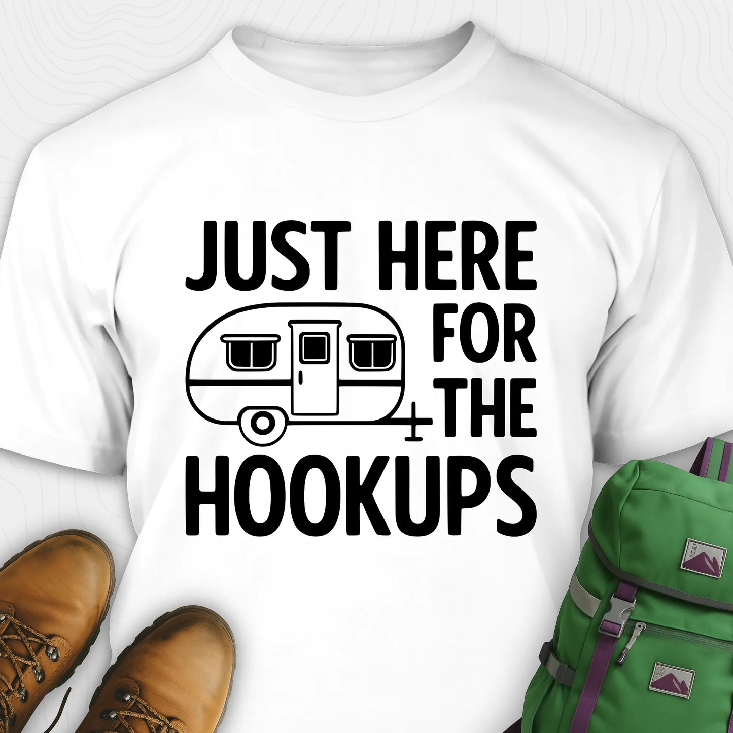 White hook ups t-shirt with trailer graphic and funny camping quote, paired with brown boots and a green backpack on a white background.
