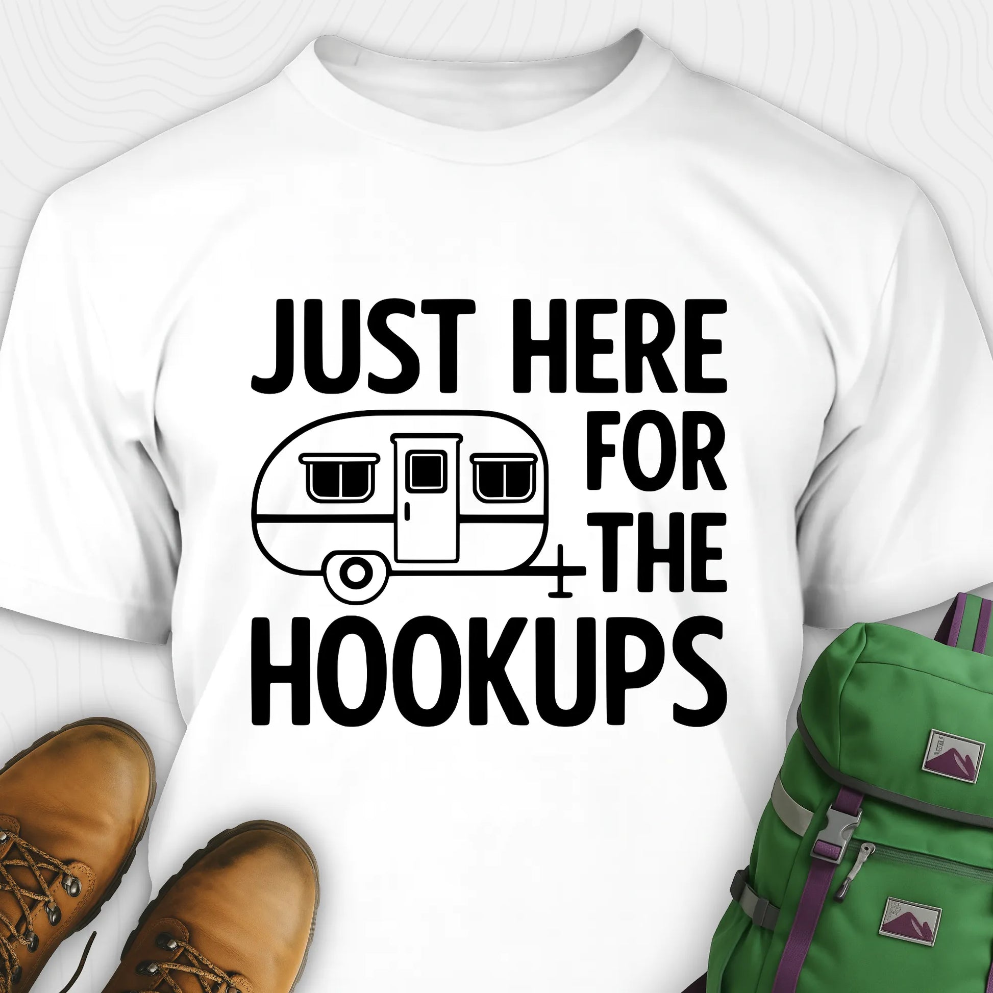 White hook ups t-shirt with trailer graphic and funny camping quote, paired with brown boots and a green backpack on a white background.