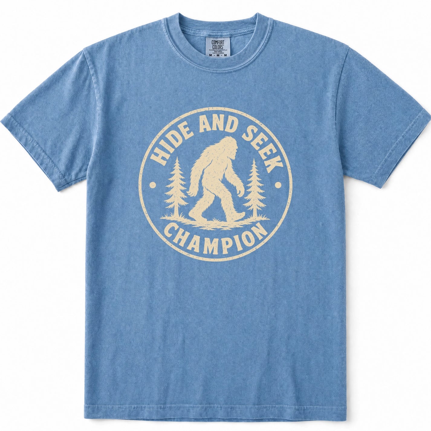 Blue Jean short-sleeve t-shirt with a cream-colored circular graphic on the chest featuring a Bigfoot silhouette and the text 'HIDE AND SEEK CHAMPION' with pine trees.