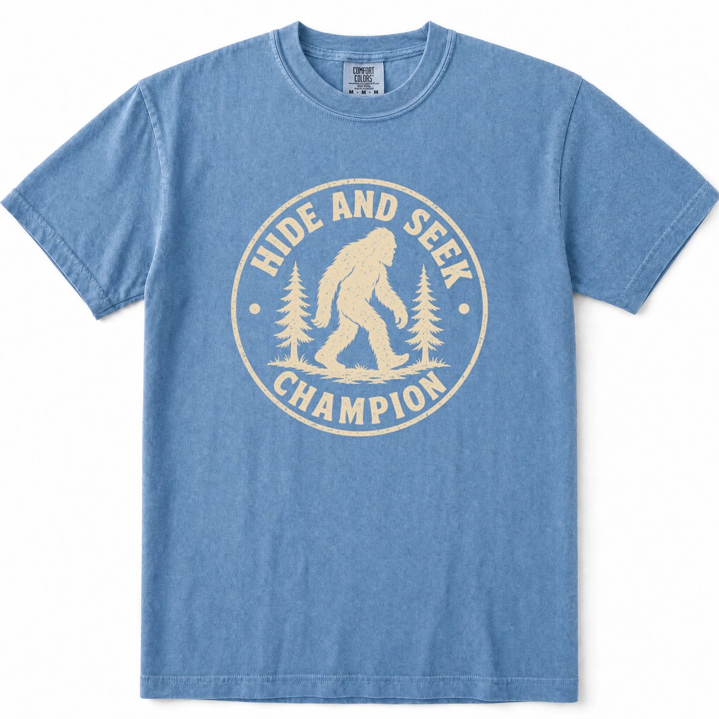 Blue Jean short-sleeve t-shirt with a cream-colored circular graphic on the chest featuring a Bigfoot silhouette and the text 'HIDE AND SEEK CHAMPION' with pine trees.