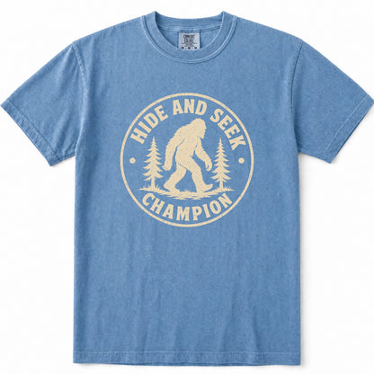 Blue Jean short-sleeve t-shirt with a cream-colored circular graphic on the chest featuring a Bigfoot silhouette and the text 'HIDE AND SEEK CHAMPION' with pine trees.