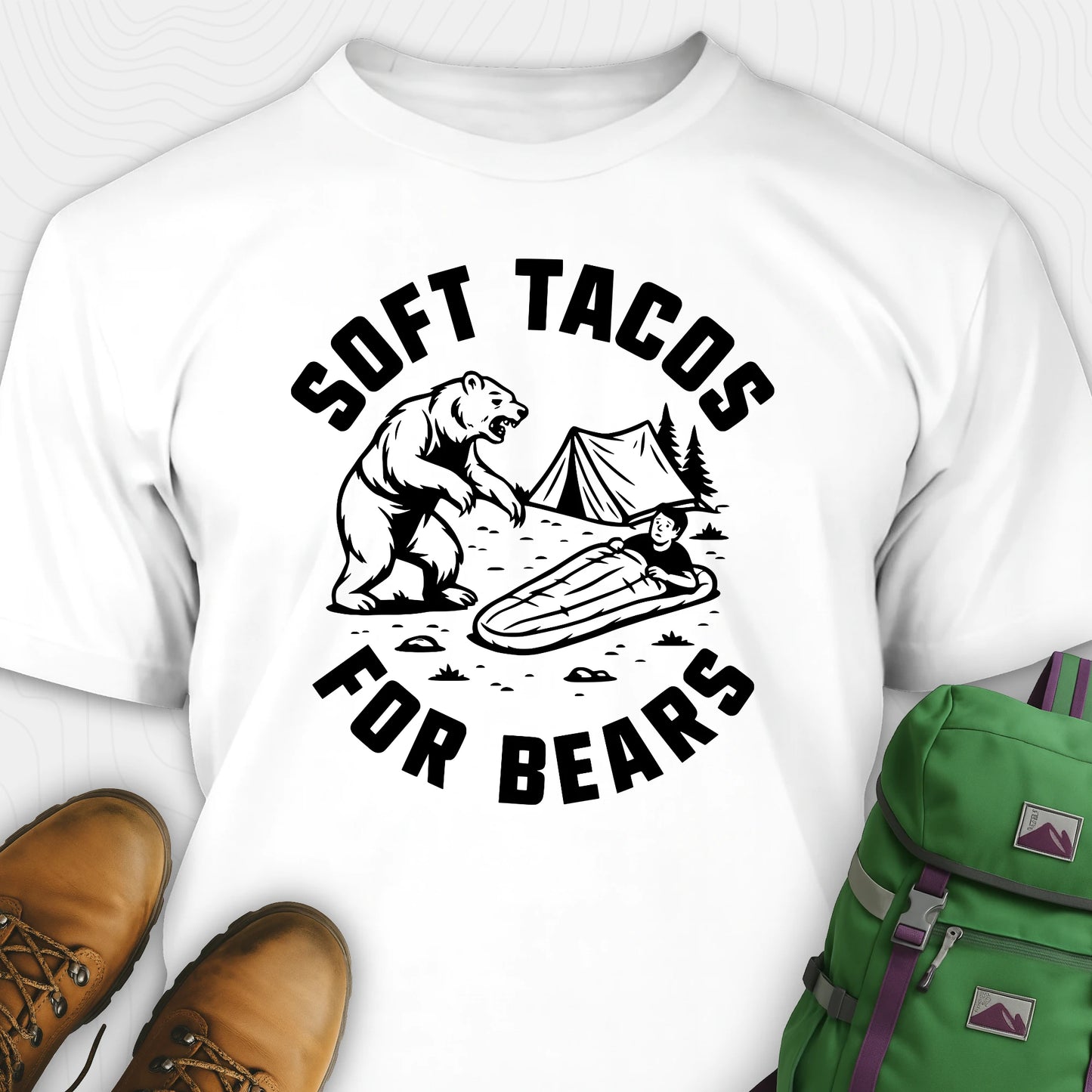 White funny bear shirt with bear graphic and text 'Soft Tacos for Bears' on a white background. surrounded by hiking boots and a green backpack.