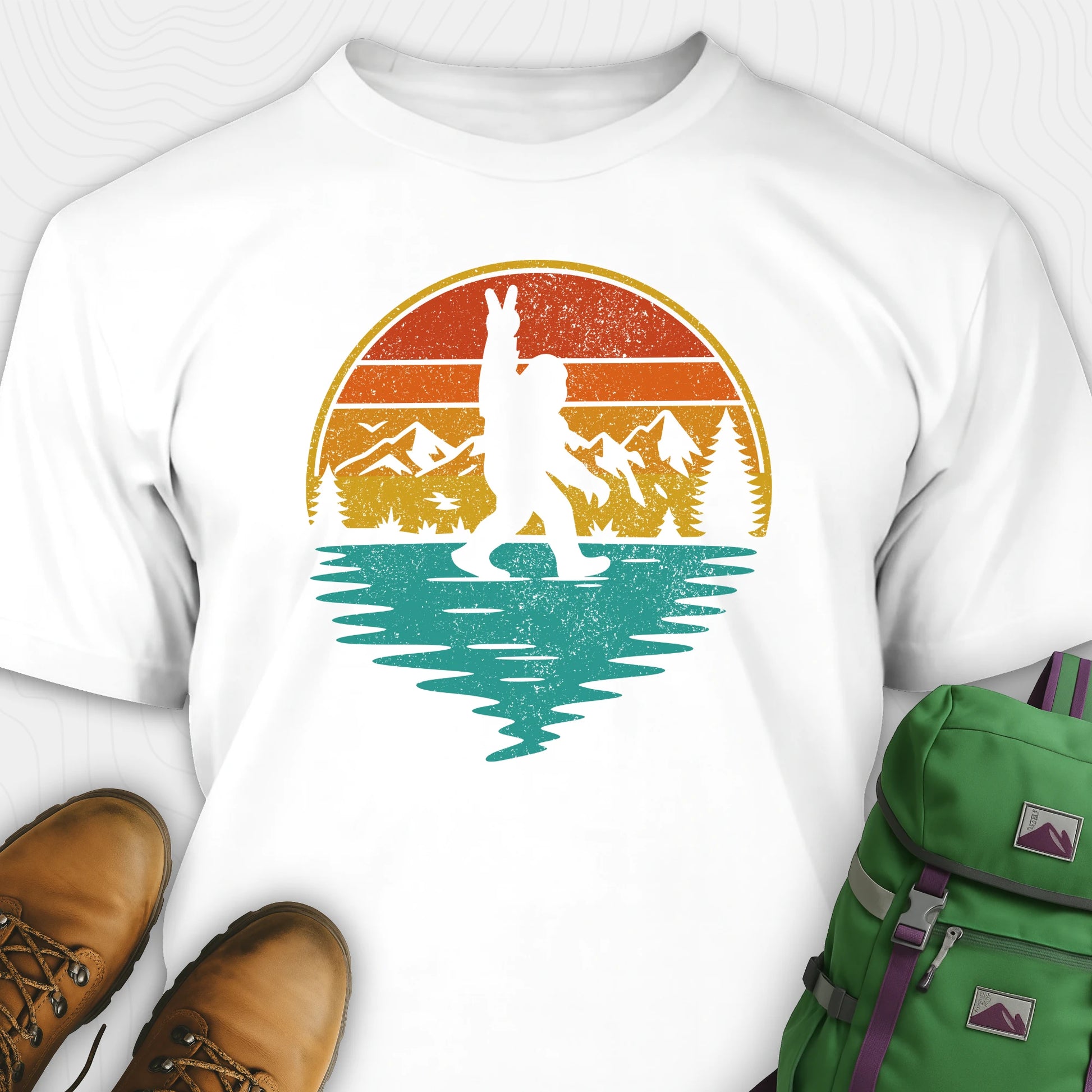 White bigfoot shirt for men with retro mountain graphic beside boots and green backpack
