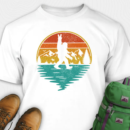 White bigfoot shirt for men with retro mountain graphic beside boots and green backpack
