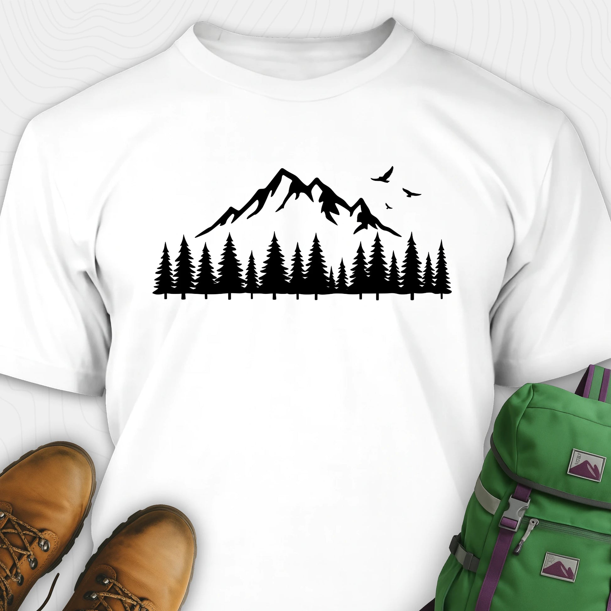 White wild t-shirt with a minimalist mountain and pine tree graphic, shown flat with boots and a backpack