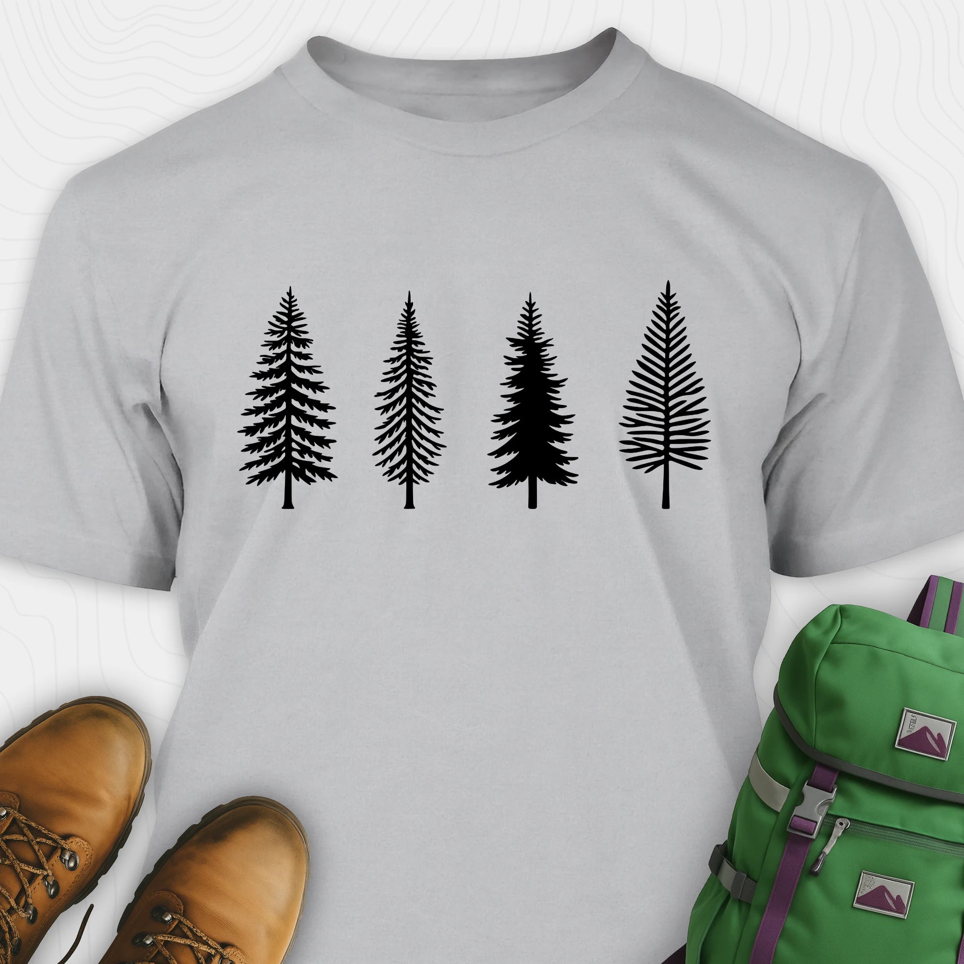 Sport grey pine tree shirt with black minimalist forest graphic, brown boots, and a green backpack on a light gray background