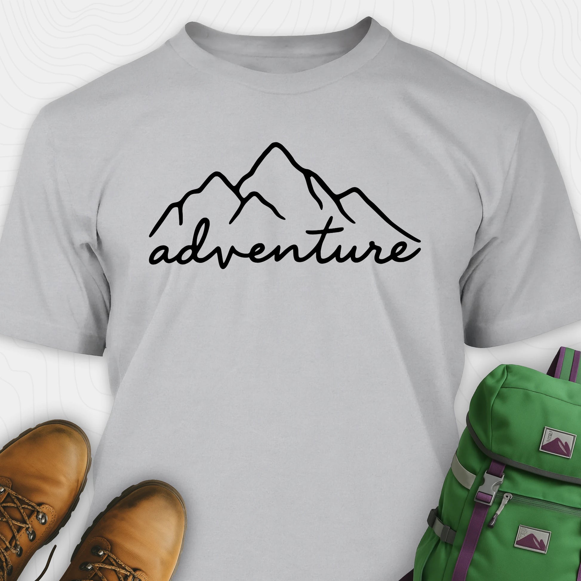 Grey adventure shirt with mountain design and 'adventure' text, paired with brown boots and a green backpack.