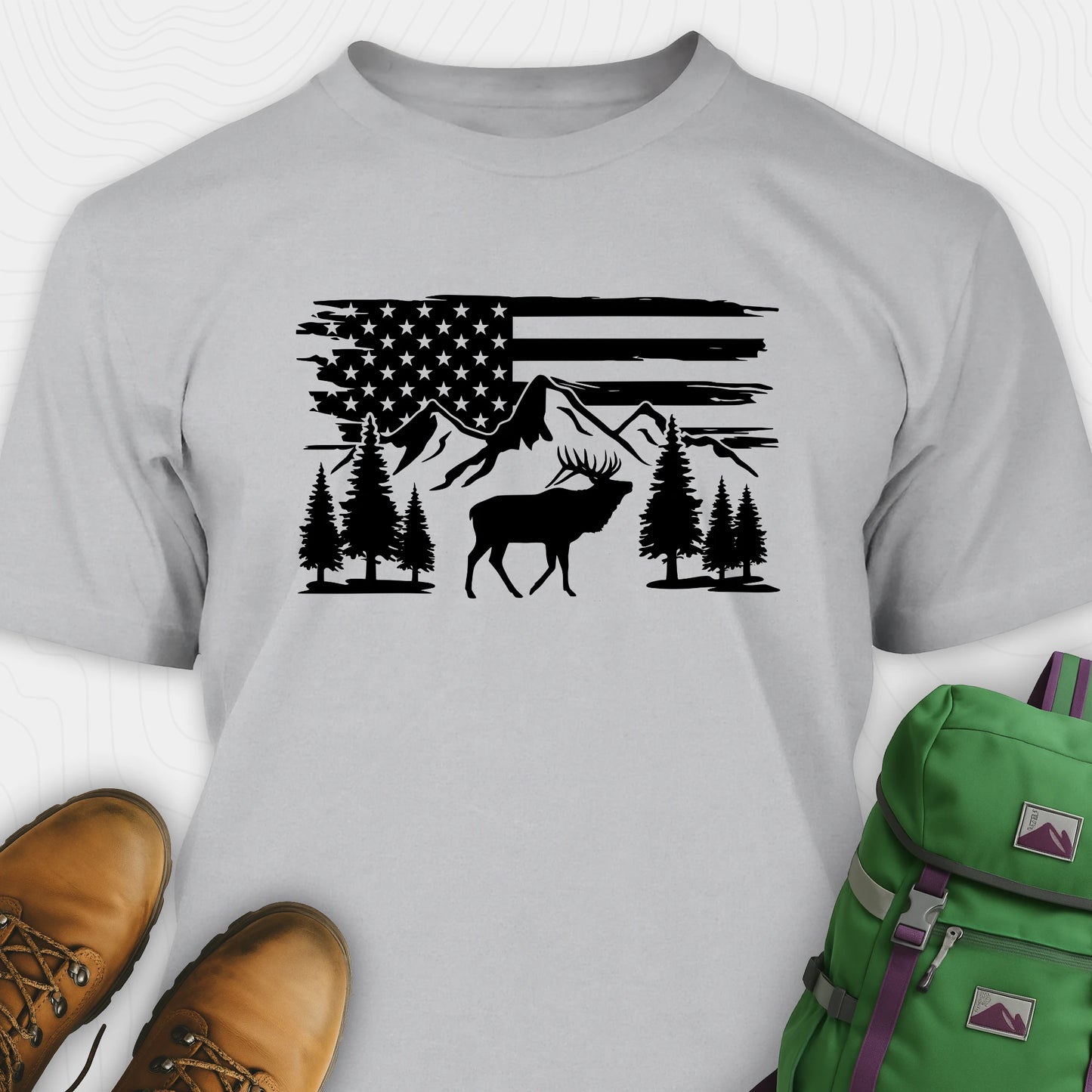 Gray deer t-shirt with black graphic design of an American flag, trees, and a moose, accompanied by brown boots and a green backpack.