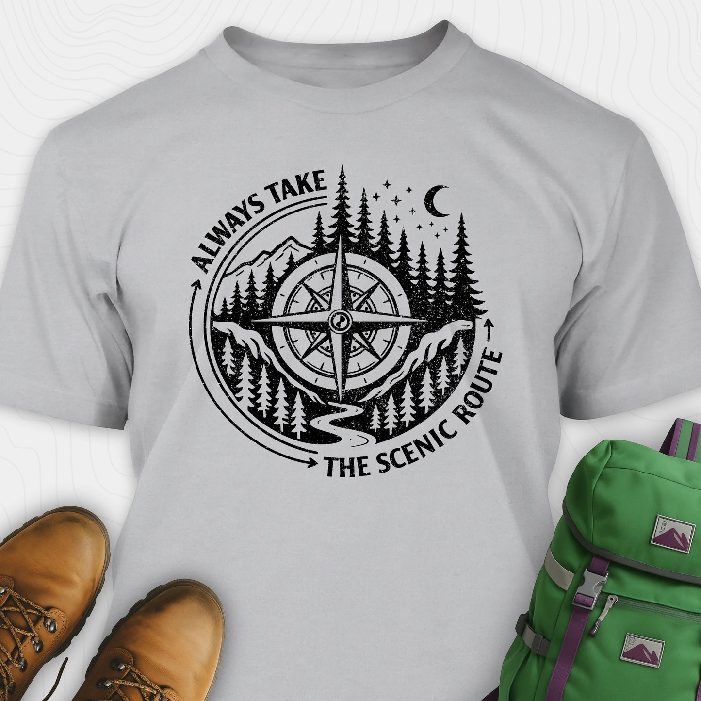 Gray always take the scenic route t-shirt with compass graphic and text, paired with hiking boots and a green backpack.