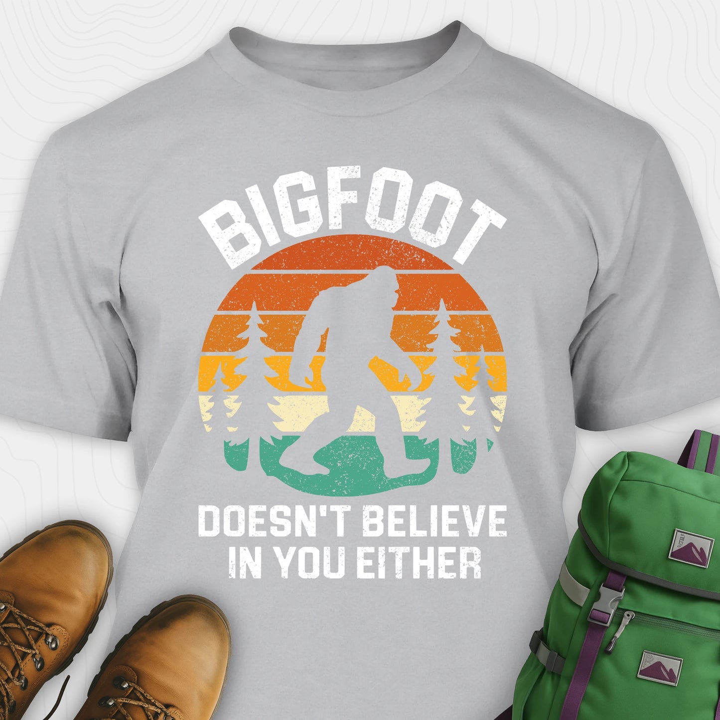 Grey funny Bigfoot shirt with sasquatch silhouette and humorous saying, paired with brown boots and a green backpack on a white background.
