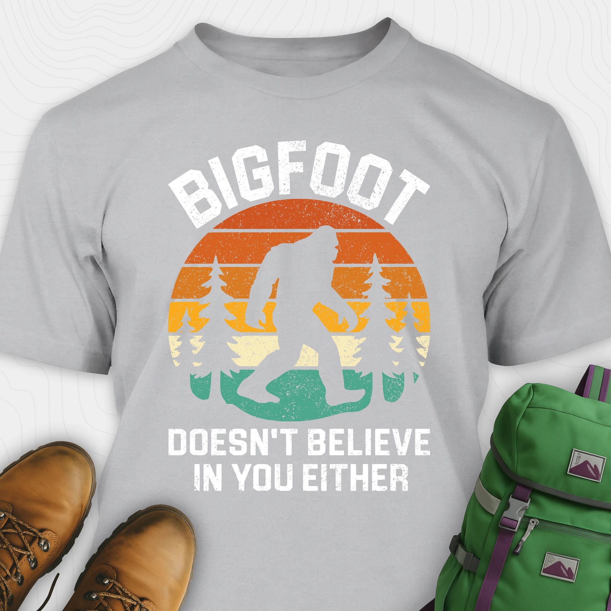 Grey funny Bigfoot shirt with sasquatch silhouette and humorous saying, paired with brown boots and a green backpack on a white background.