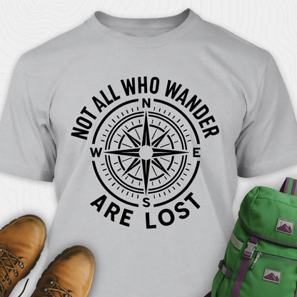 Sport grey Not All Who Wander Are Lost T-shirt with compass design and text, paired with brown boots and a green backpack on a white background.