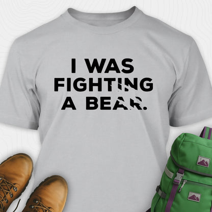 Grey I was fighting a bear shirt with a bear scratch mark graphic, paired with brown boots and a green backpack on a white background.