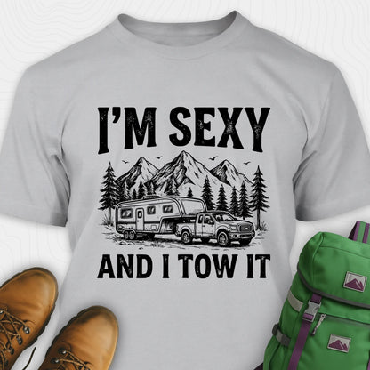 Gray t-shirt with 'I'm Sexy and I Tow It' graphic design, brown boots, and green backpack on a white background