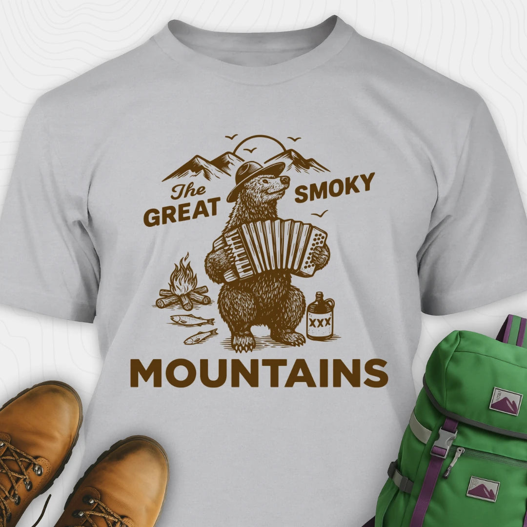 Gray t-shirt with bear playing accordion graphic and 'The Great Smoky Mountains' text, surrounded by hiking boots and a green backpack.