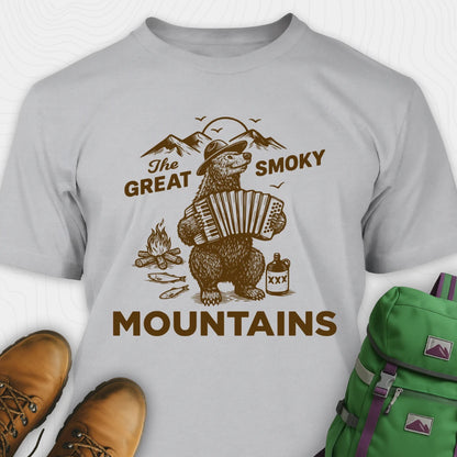 Gray t-shirt with bear playing accordion graphic and 'The Great Smoky Mountains' text, surrounded by hiking boots and a green backpack.