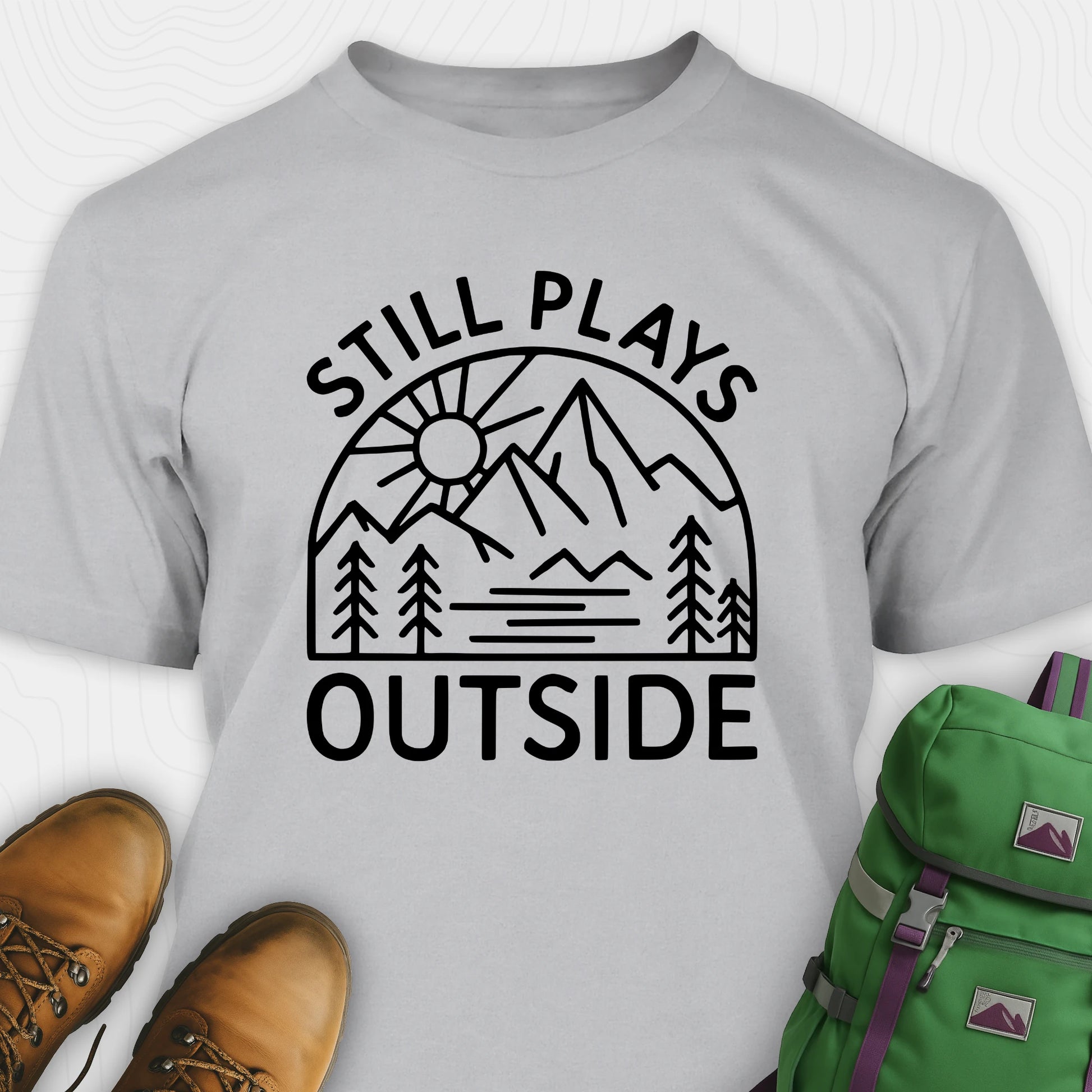 Gray t-shirt with 'Still Plays Outside' graphic design, paired with brown boots and a green backpack on a light gray background.