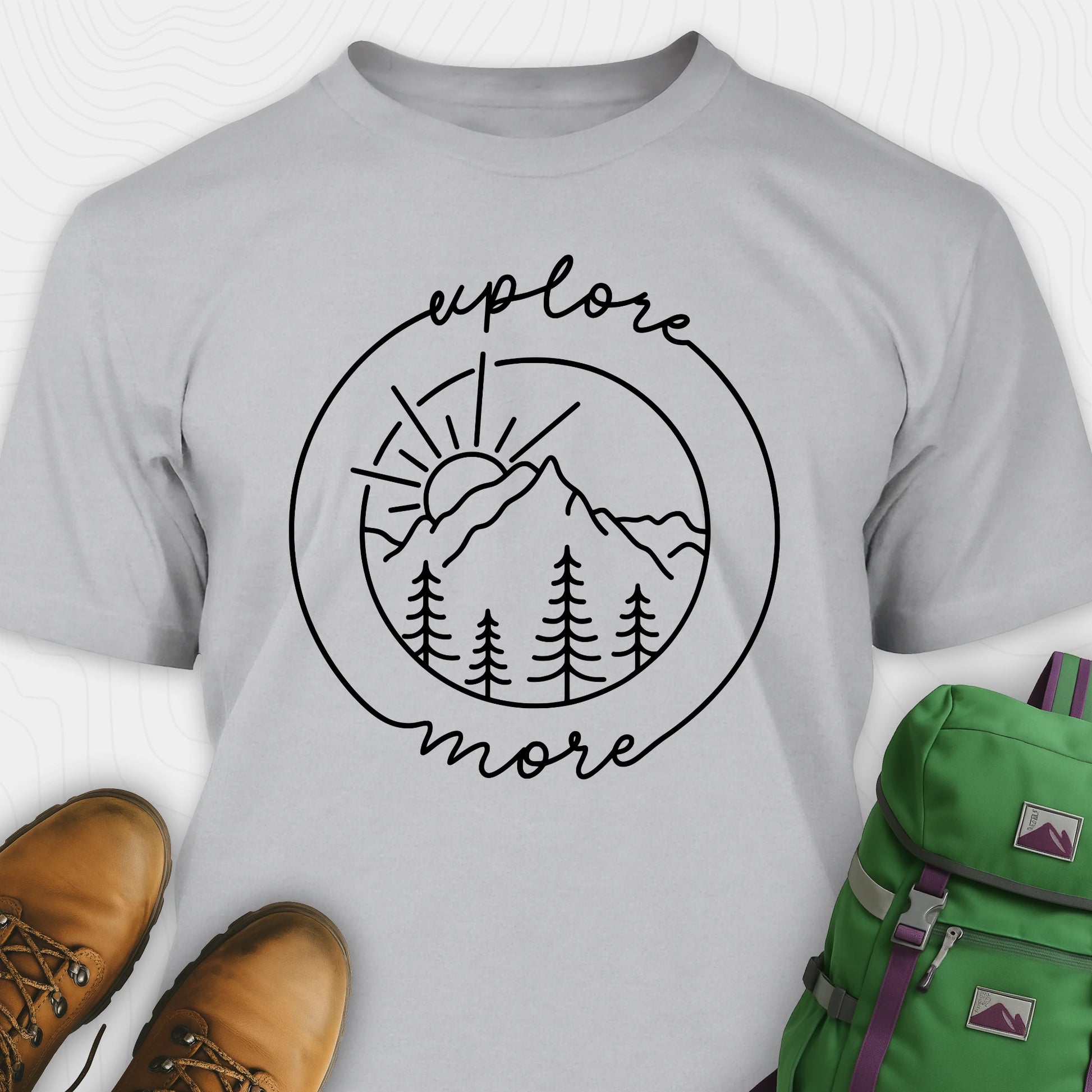 Grey explorer shirt with 'explore more' graphic design, brown boots, and green backpack on a white background