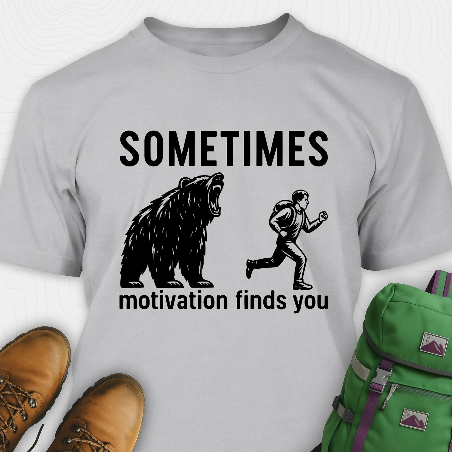 Grey motivational t-shirt with a bear and person graphic and text, paired with brown boots and a green backpack on a white background.