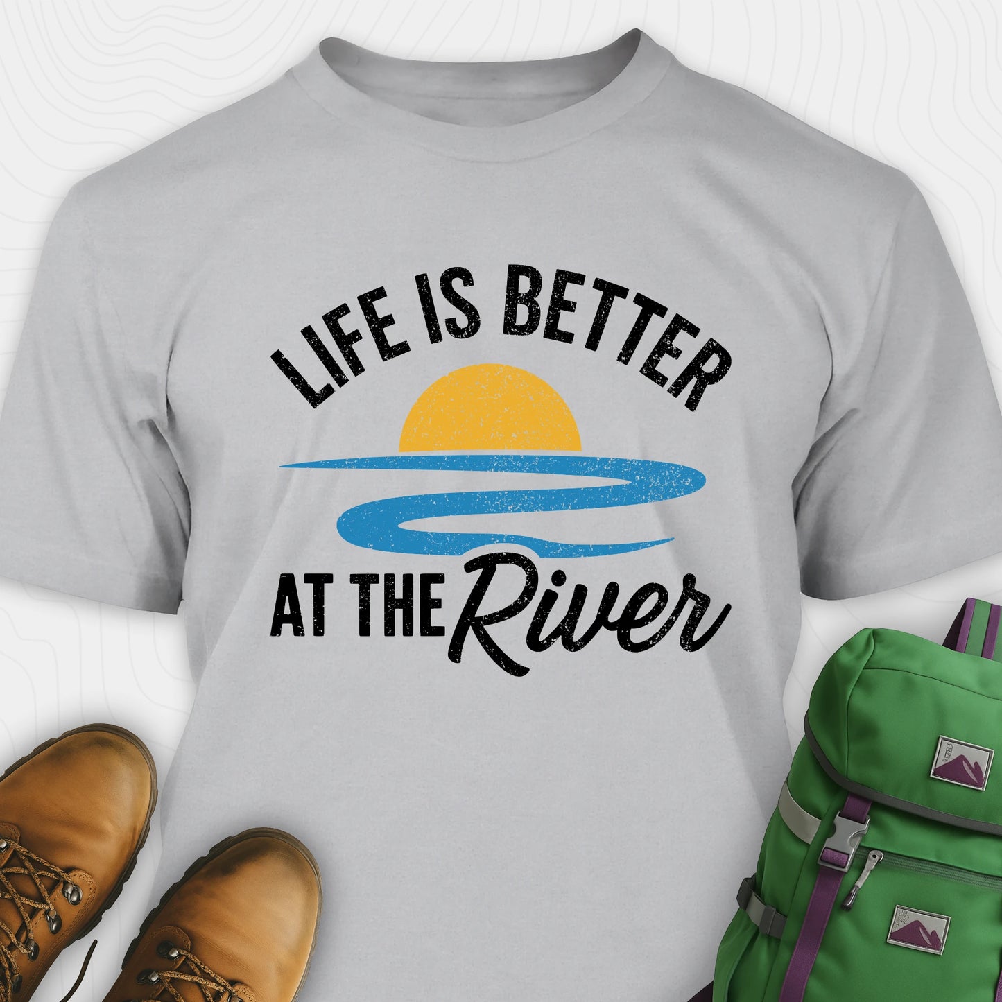 Gray river shirt with 'Life is Better at the River' design, paired with brown boots and a green backpack on a light gray background.