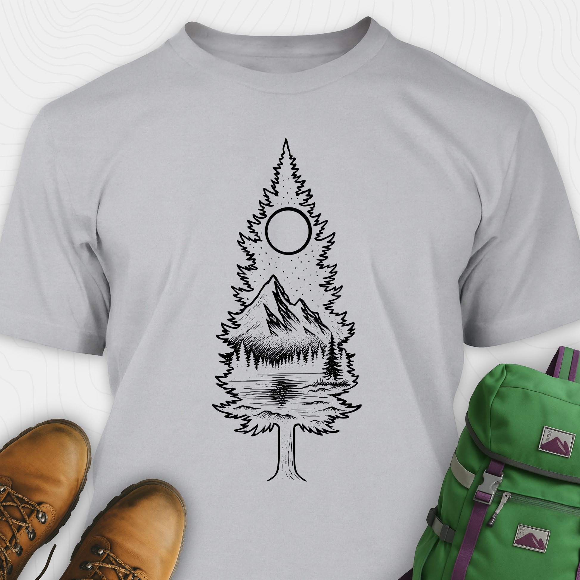 A gray tree t-shirt featuring a detailed white graphic of a pine tree silhouette. The tree outline contains a landscape scene: a mountain peak rising over a forested shoreline, with its reflection visible in a lake below. Above the mountain is a starry night sky and a full or partial circle representing the moon or sun. The t-shirt is displayed flat with brown leather boots to the lower left and a green and purple backpack to the lower right, suggesting an outdoor or hiking theme.