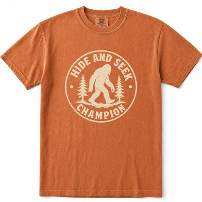 Yam short-sleeve crewneck t-shirt featuring a distressed cream-colored circular graphic on the chest with a Bigfoot silhouette and the words 'HIDE AND SEEK CHAMPION' alongside pine trees.