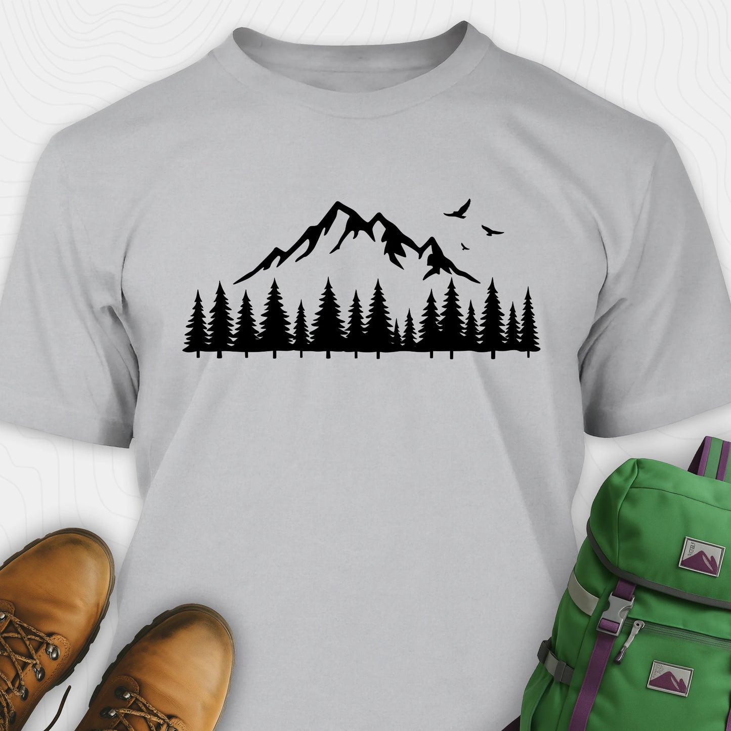 Grey wild t-shirt with a minimalist mountain and pine tree graphic, shown flat with boots and a backpack