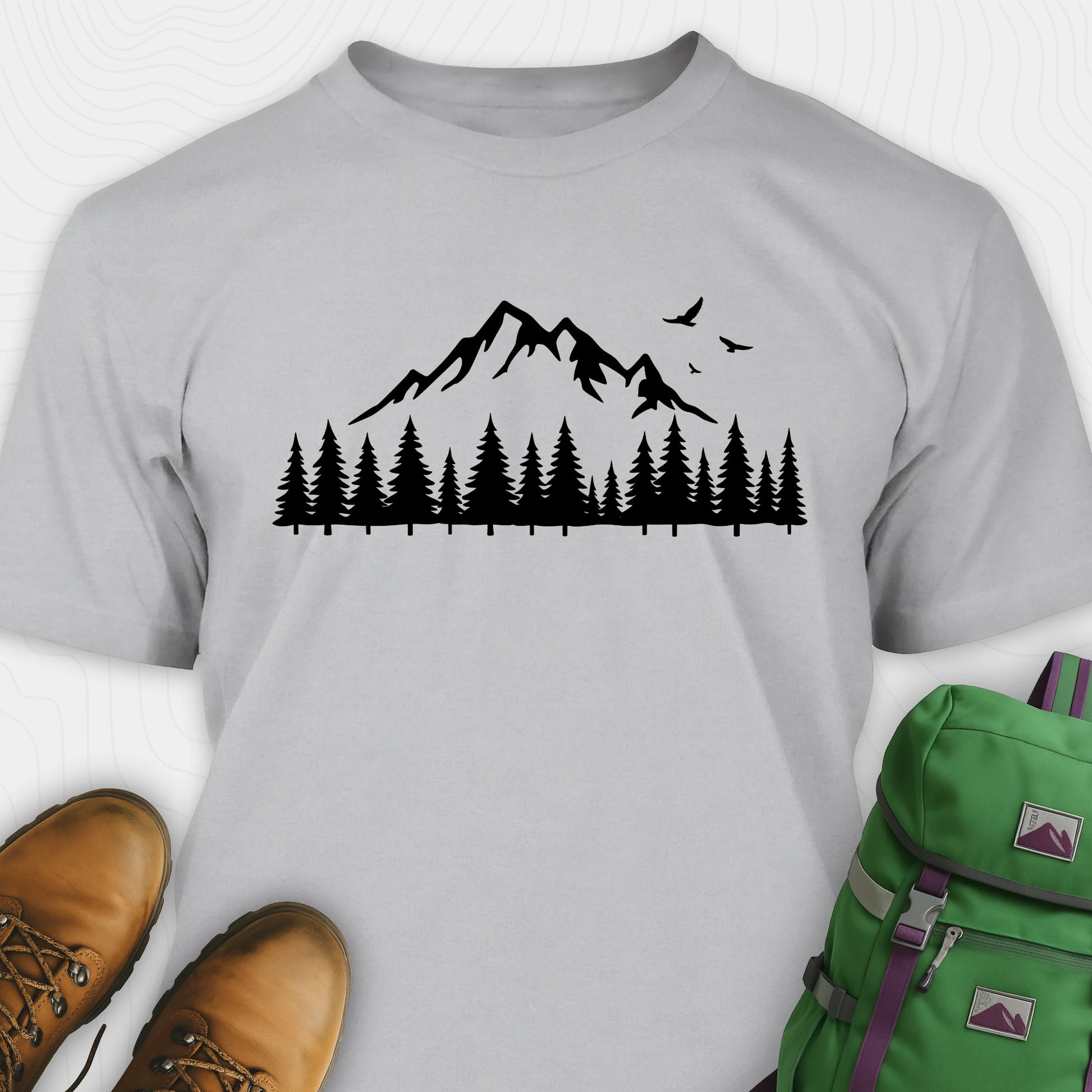 Grey wild t-shirt with a minimalist mountain and pine tree graphic, shown flat with boots and a backpack