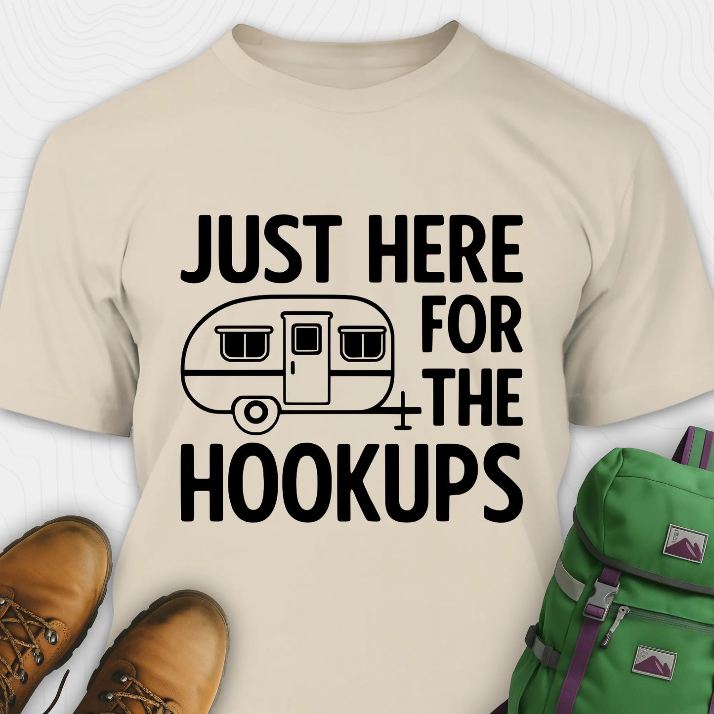 Sand hook ups t-shirt with trailer graphic and funny camping quote, paired with brown boots and a green backpack on a white background.