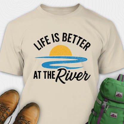 Sand river shirt with 'Life is Better at the River' design, paired with brown boots and a green backpack on a light gray background.