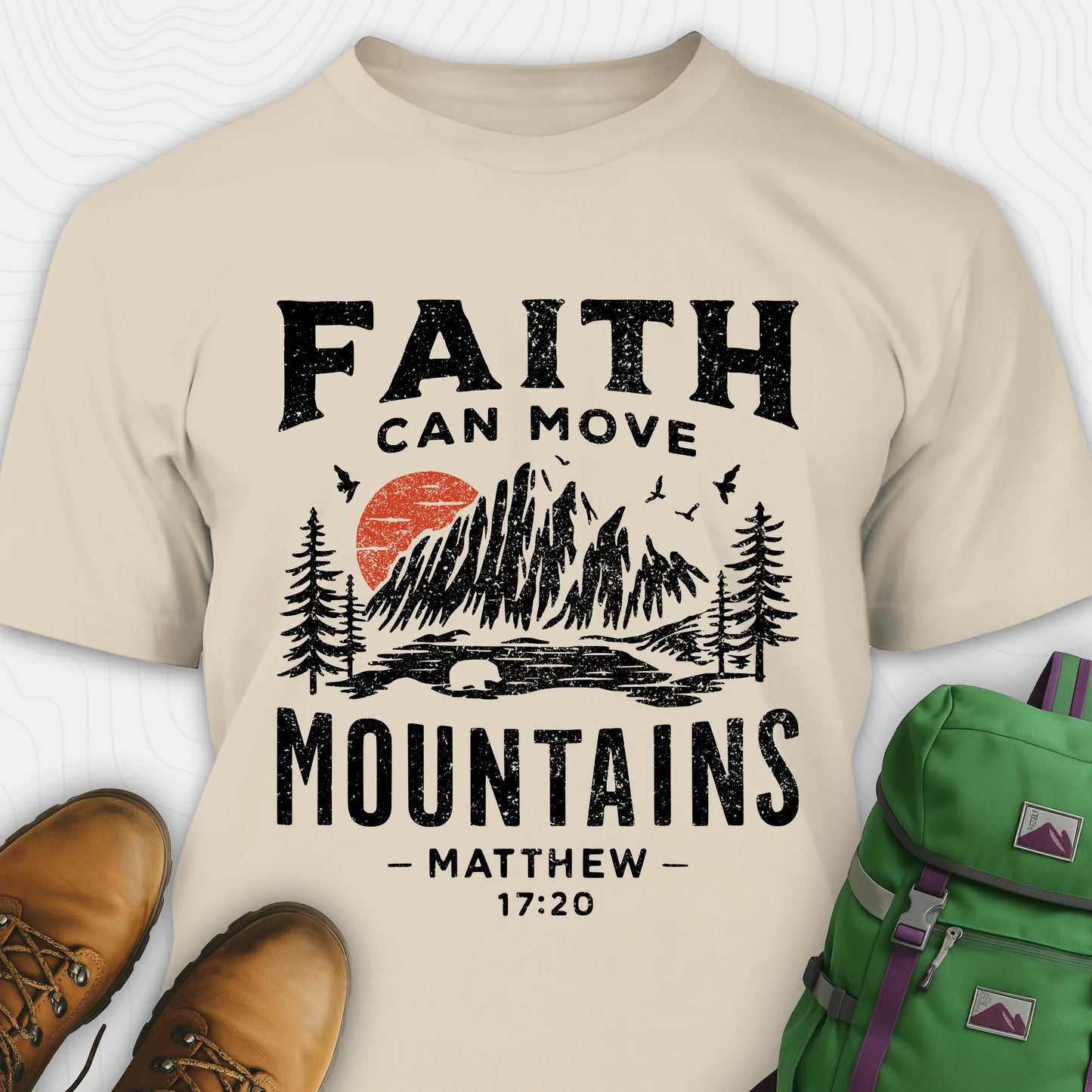 Sand bible verse t-shirt with 'Faith can move mountains' design and hiking boots and backpack on a white background