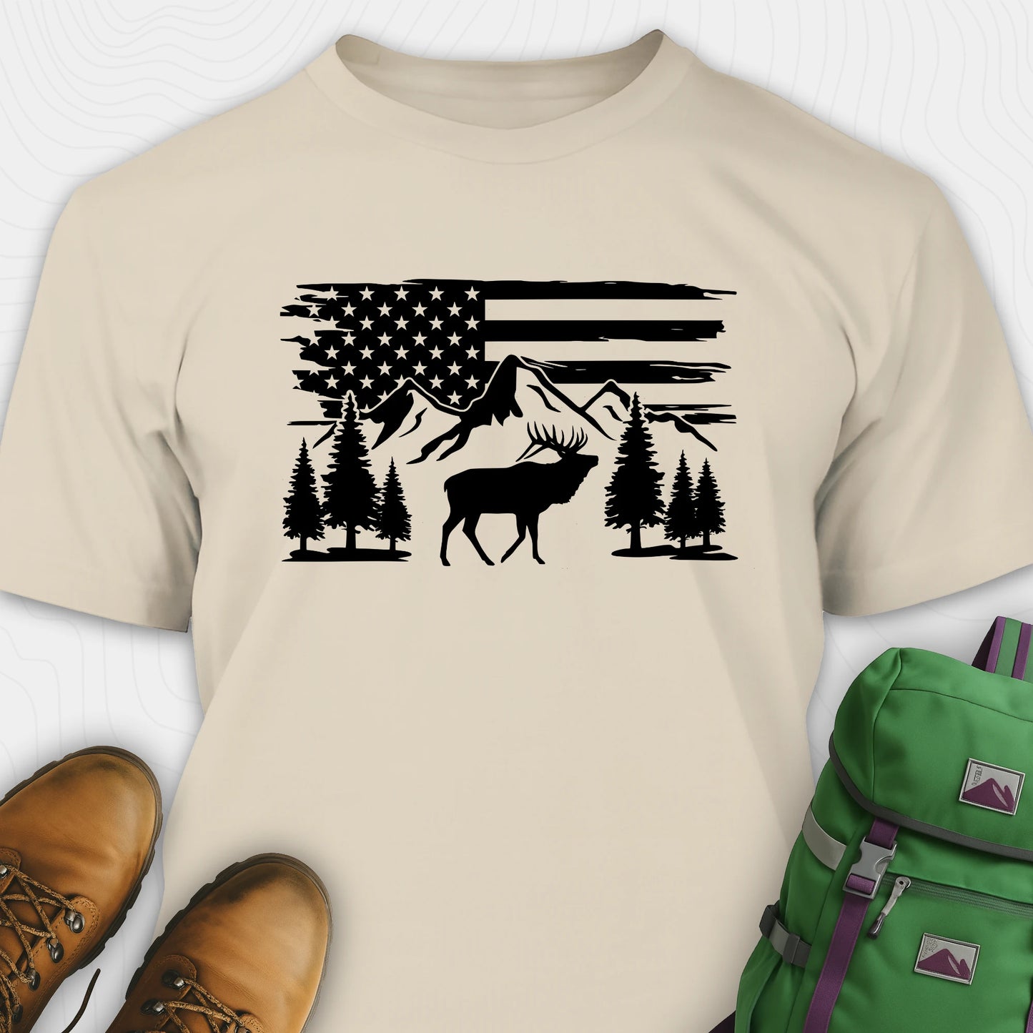 Beige deer t-shirt with black graphic design of an American flag, trees, and a moose, accompanied by brown boots and a green backpack.