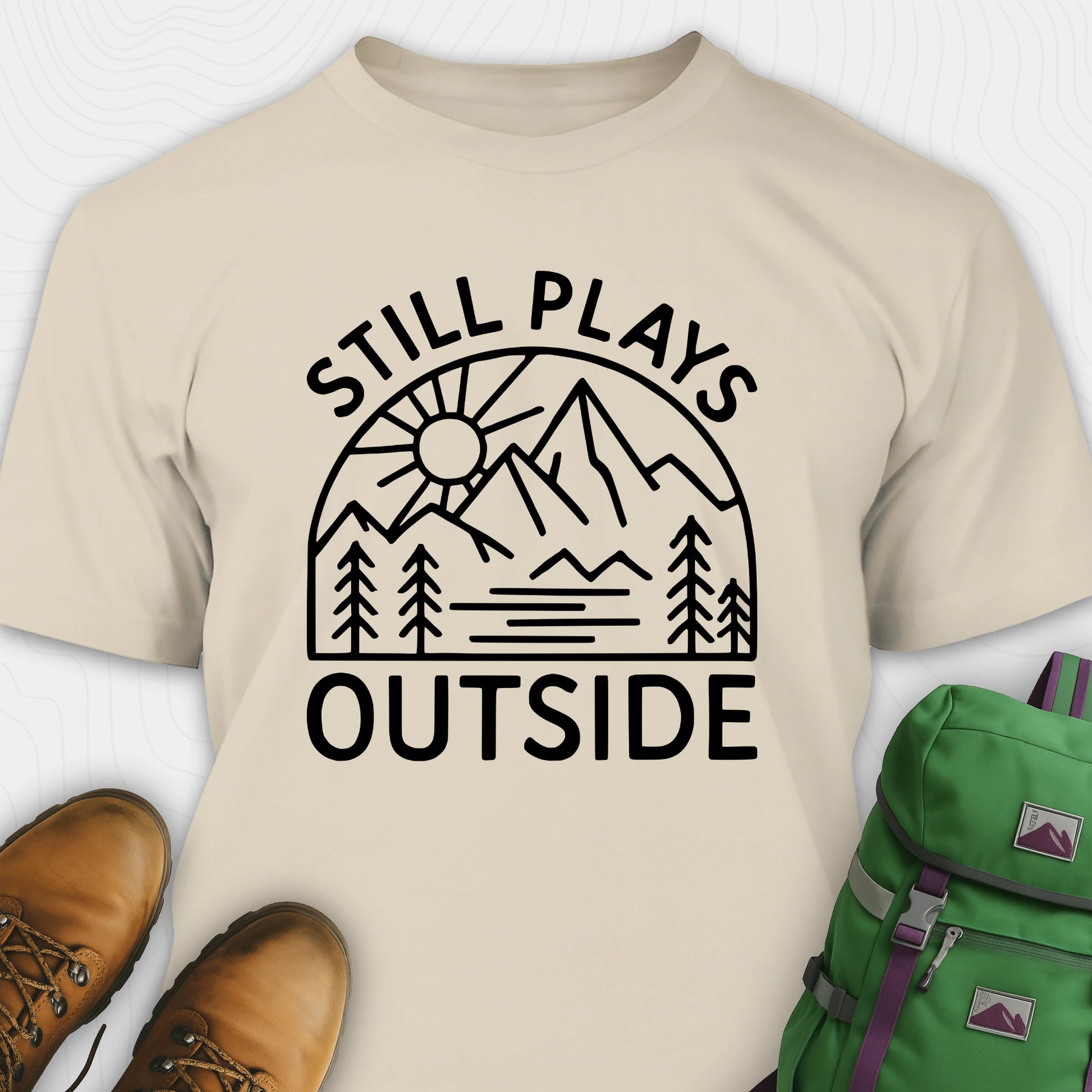 Natural t-shirt with 'Still Plays Outside' graphic design, paired with brown boots and a green backpack on a light gray background.