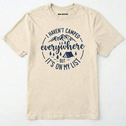 Natural funny camping shirt saying with mountains, trees, tent graphic and "I Haven't Camped Everywhere But It's On My List" text on a white background.