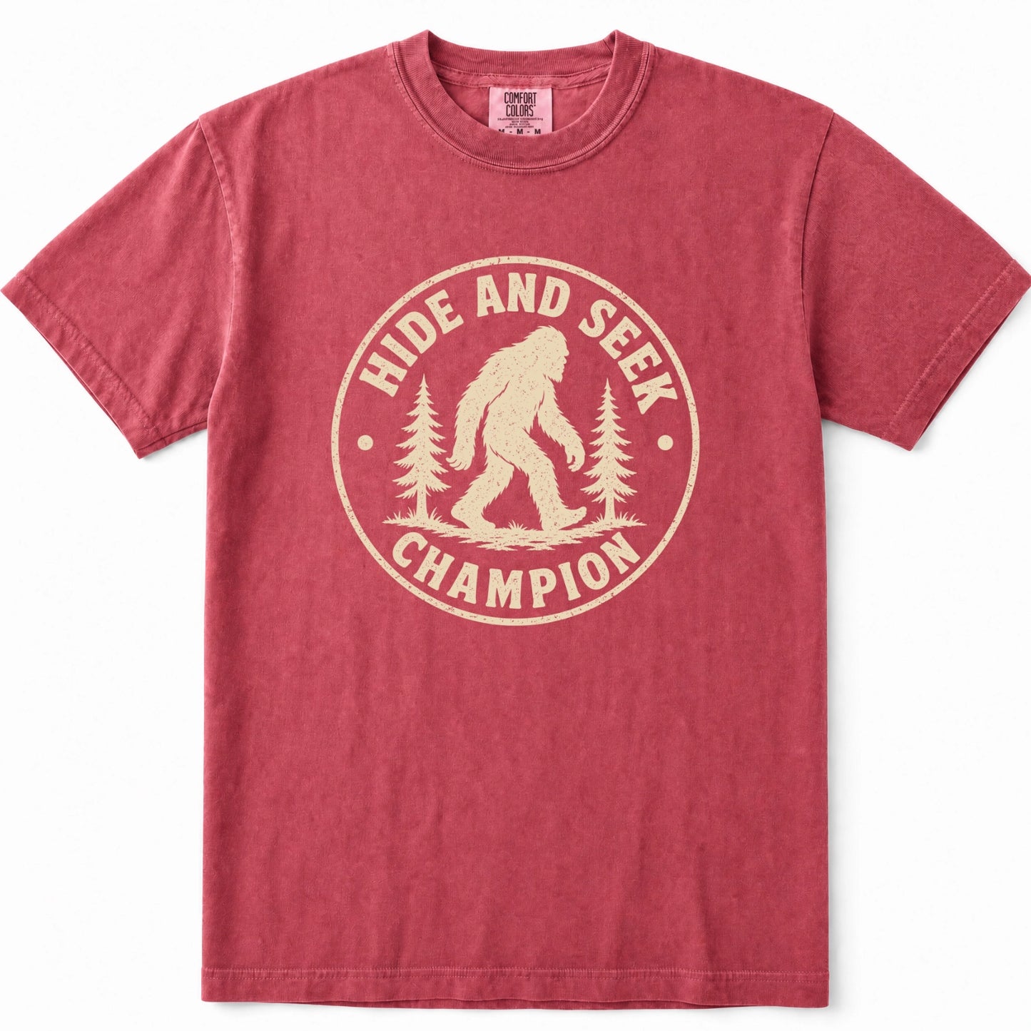 Crimson short-sleeve crewneck t-shirt in a washed, heathered red color, featuring a vintage-style beige graphic on the chest with a Bigfoot silhouette, pine trees, and the text 'HIDE AND SEEK CHAMPION'.