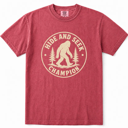 Crimson short-sleeve crewneck t-shirt in a washed, heathered red color, featuring a vintage-style beige graphic on the chest with a Bigfoot silhouette, pine trees, and the text 'HIDE AND SEEK CHAMPION'.