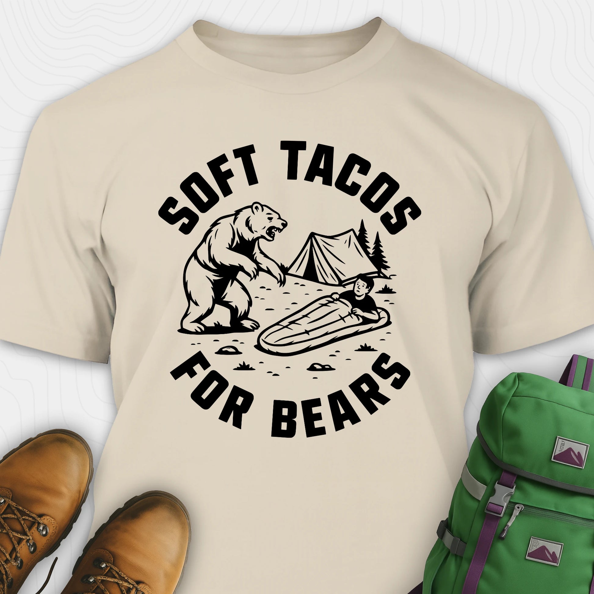 Beige funny bear shirt with bear graphic and text 'Soft Tacos for Bears' on a white background. surrounded by hiking boots and a green backpack.
